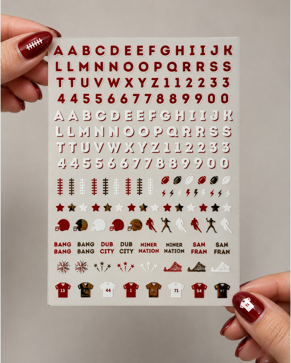 A sheet of nail stickers held by two hands with red nail polish. The sheet features the San Francisco 49ers colors (red, white, and brown) with various lettering, numbers, footballs, helmets, and team logos.